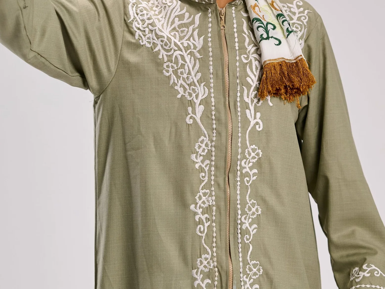 White embroidery detail on the sleeve and shoulder of a sage-green thobe under studio lighting.